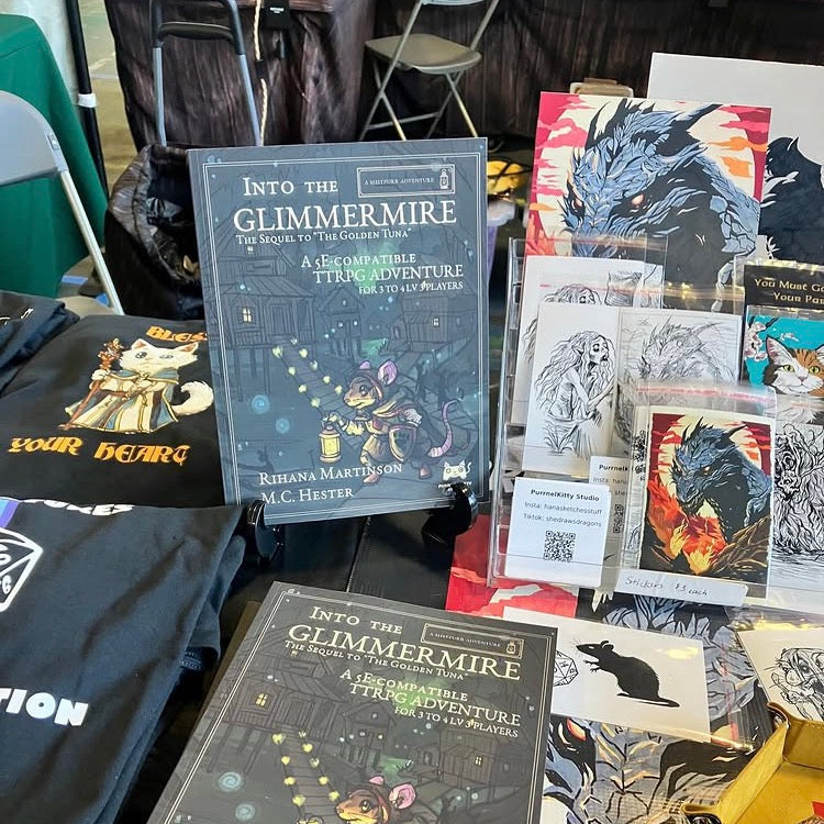 Books titled 'Into the Glimmermire' on a table with artwork and clothing items.